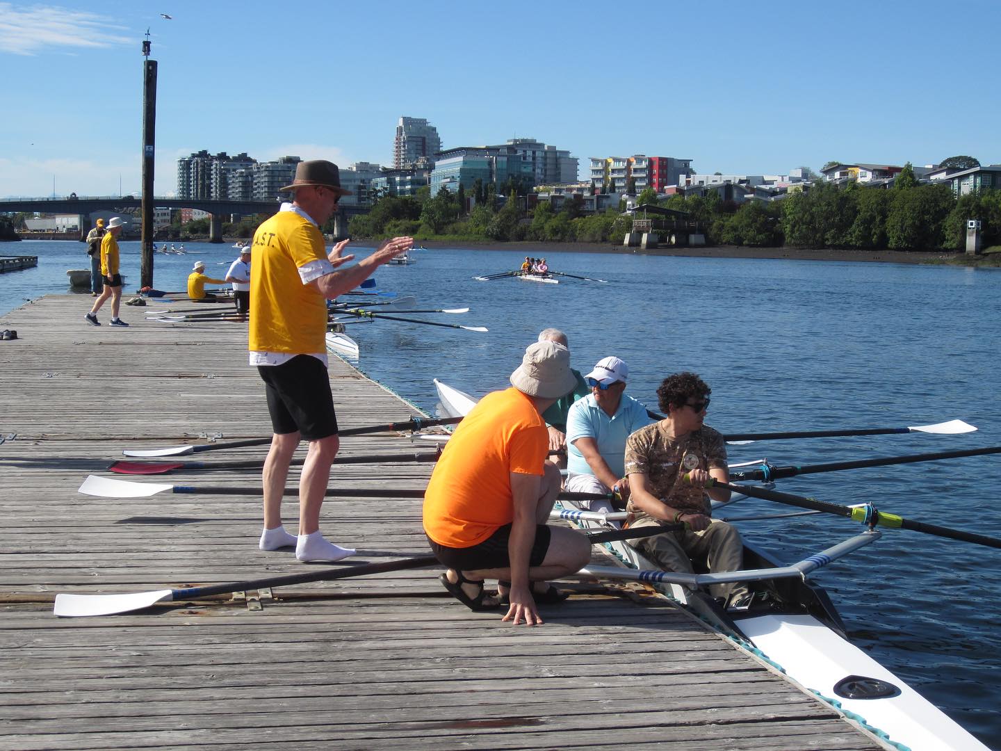 Two coaches giving instruction to rowers. The rowers are sitting in a boat at the dock. The coaches are on the dock. One is standing, the other is crouching by the boat.