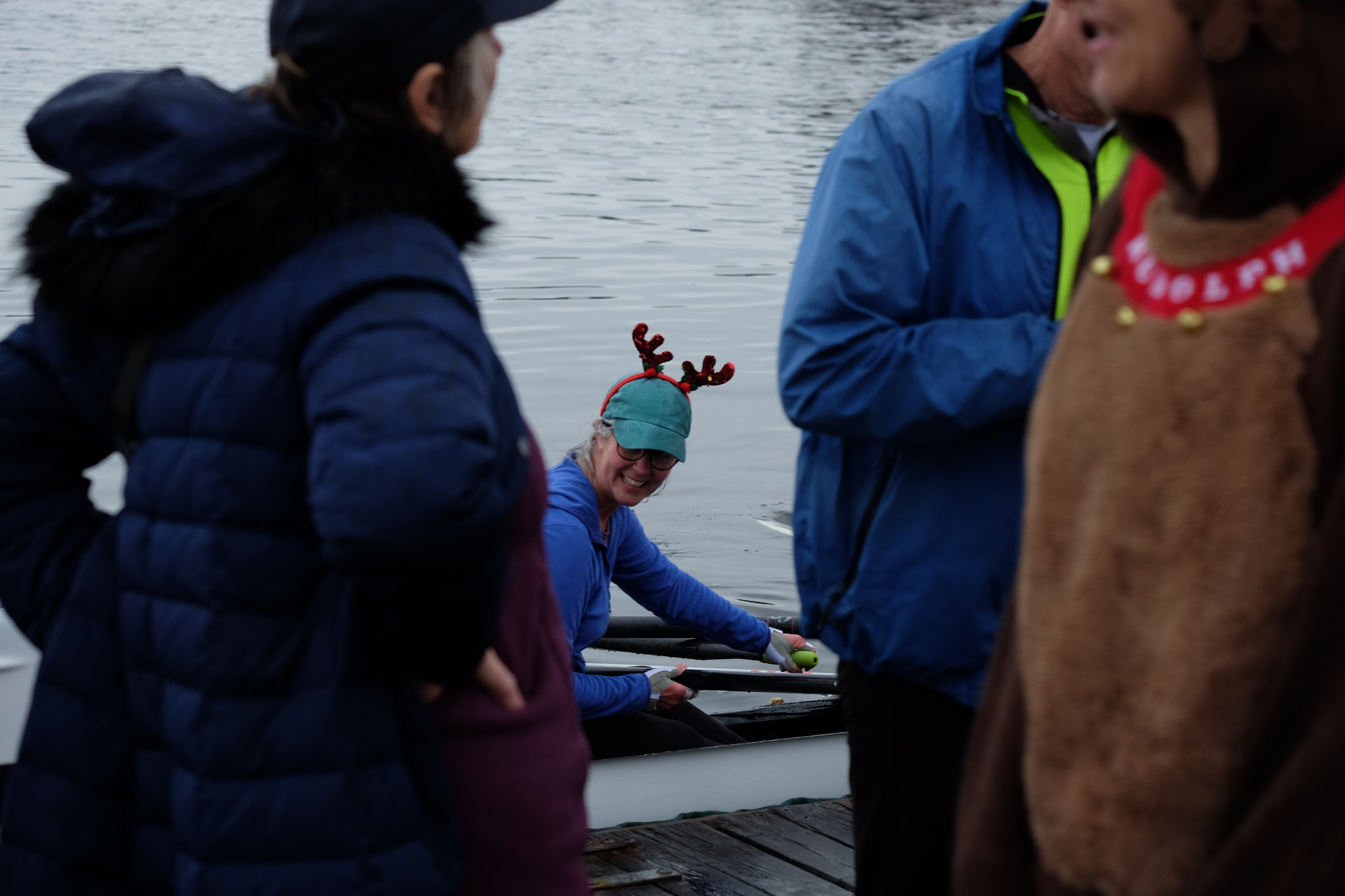 A quad docking. One rower is visible and is wearing a hat with antlers. The picture is taken through a crowd of people on the dock.