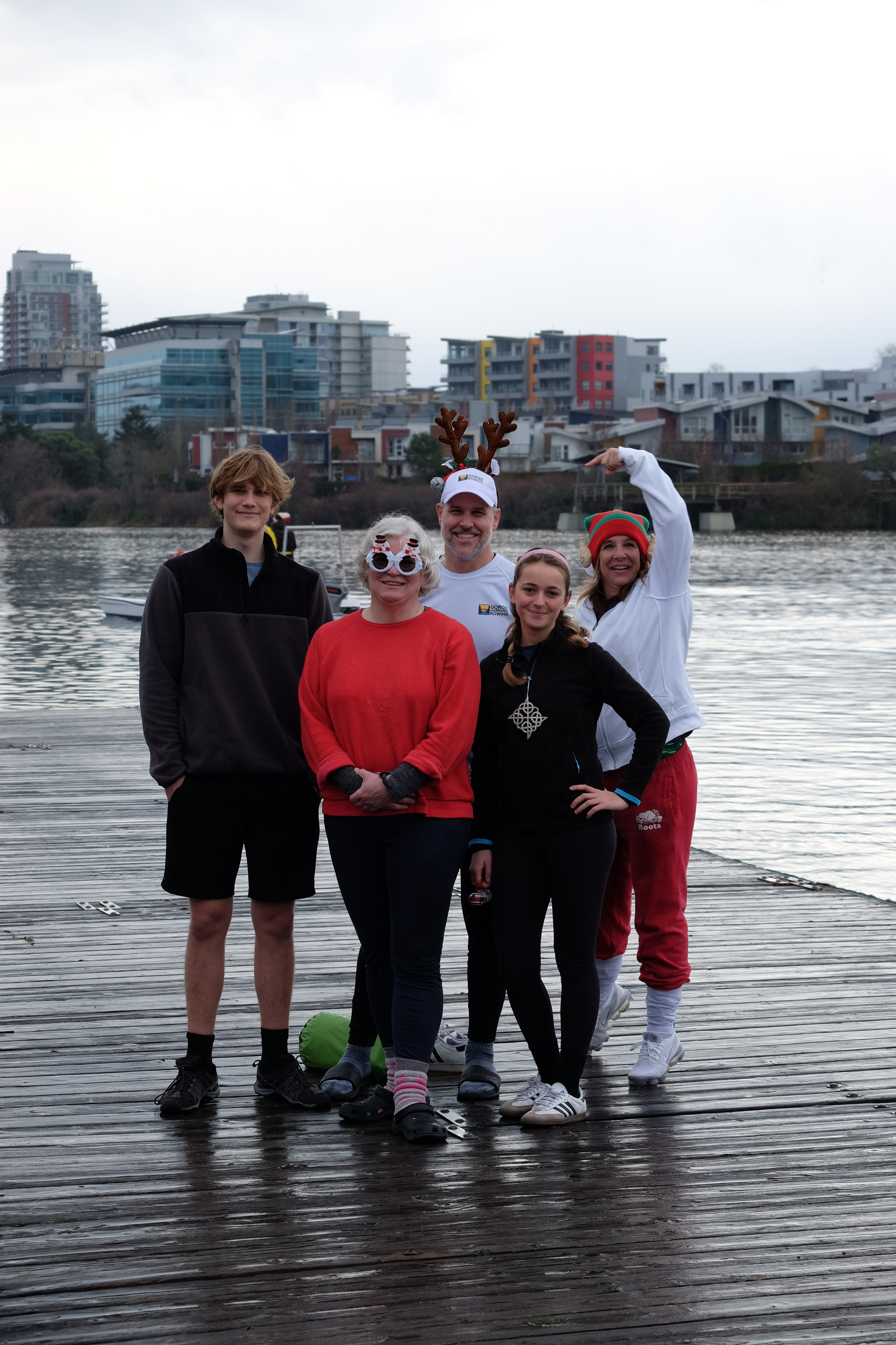 A team of four rowers standing on the dock posing for a picture. Our club manager, Rachel, has joined the picture and is pointing at one of the rowers.