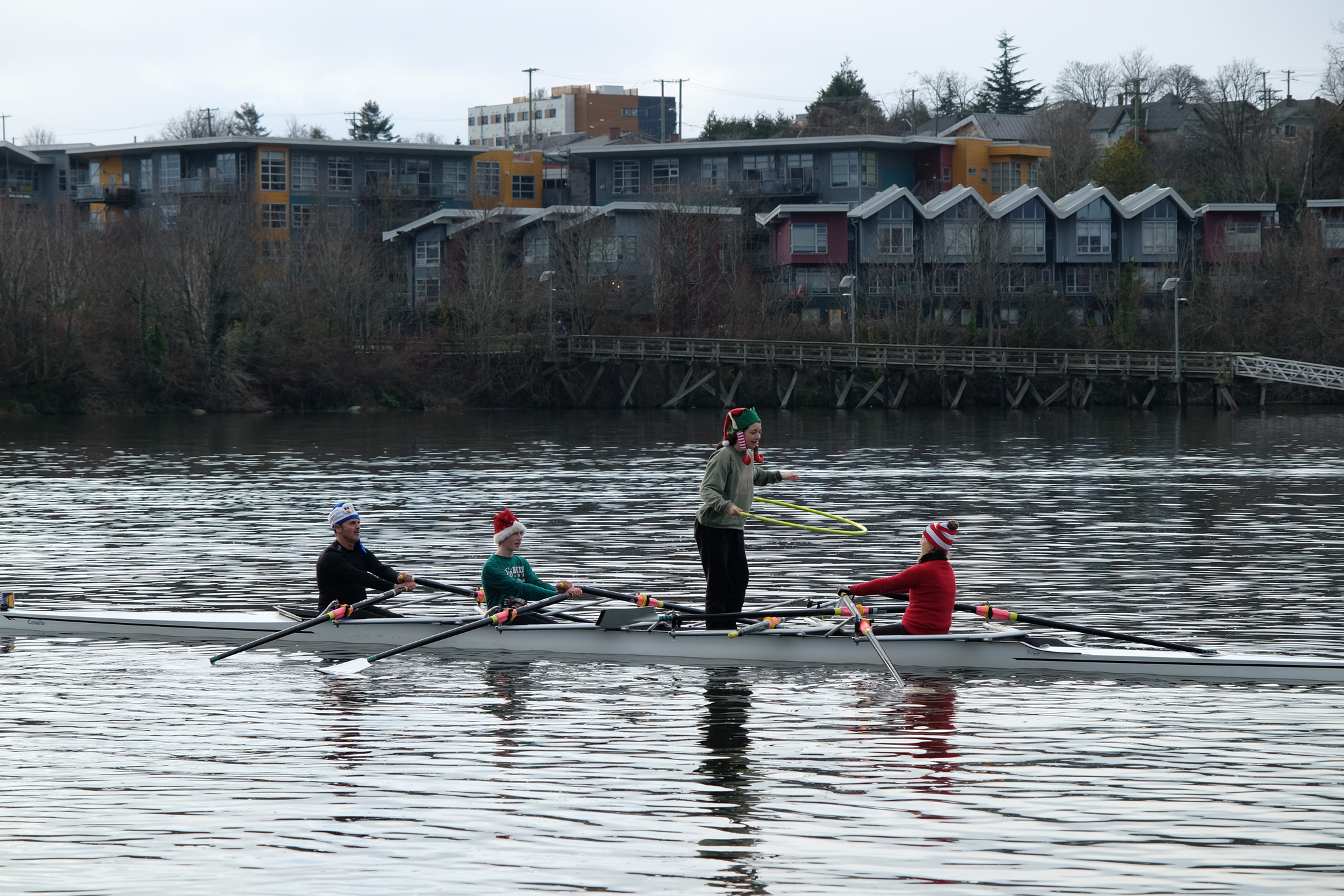 A quad on the water. One of the rowers is facing backwards. Another is standing and dancing with a hula hoop.