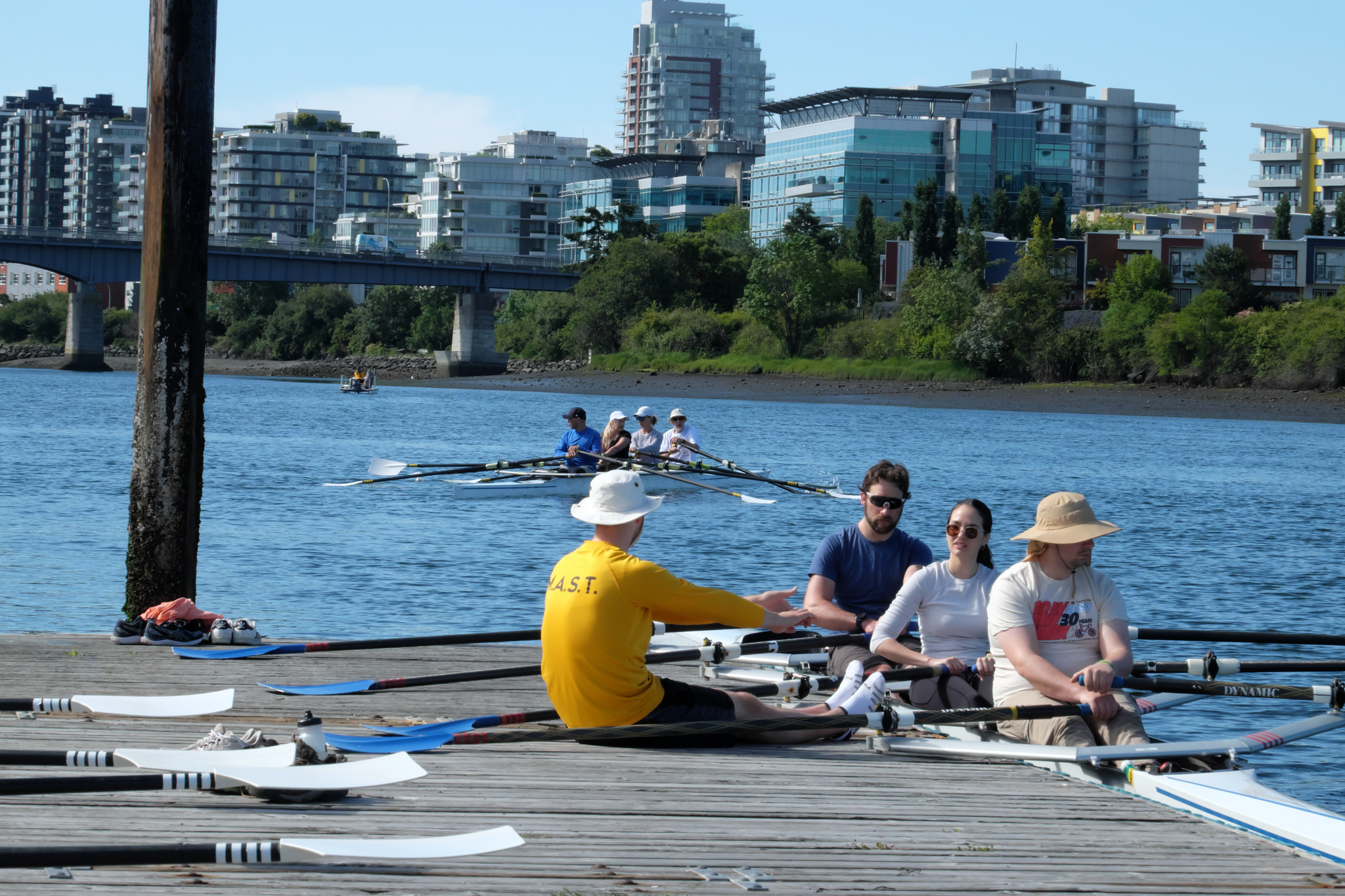 A coach giving instruction to rowers. The rowers are sitting in a boat at the dock. The coach is next to them on the dock.