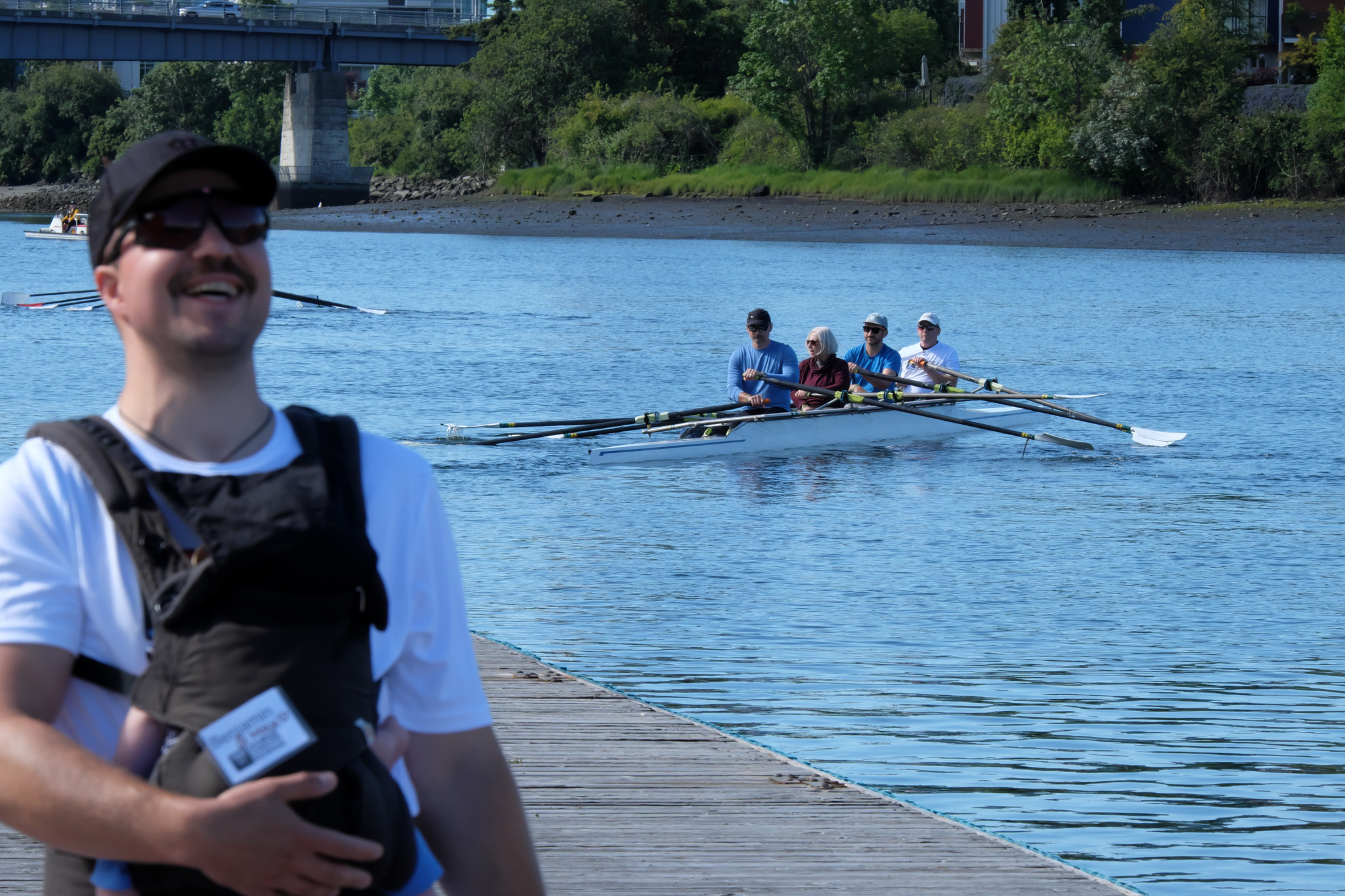 In the foreground is one of our volunteers, with their baby in a front carrier, laughing. In the background, a quad has just launched and is moving away from the dock.