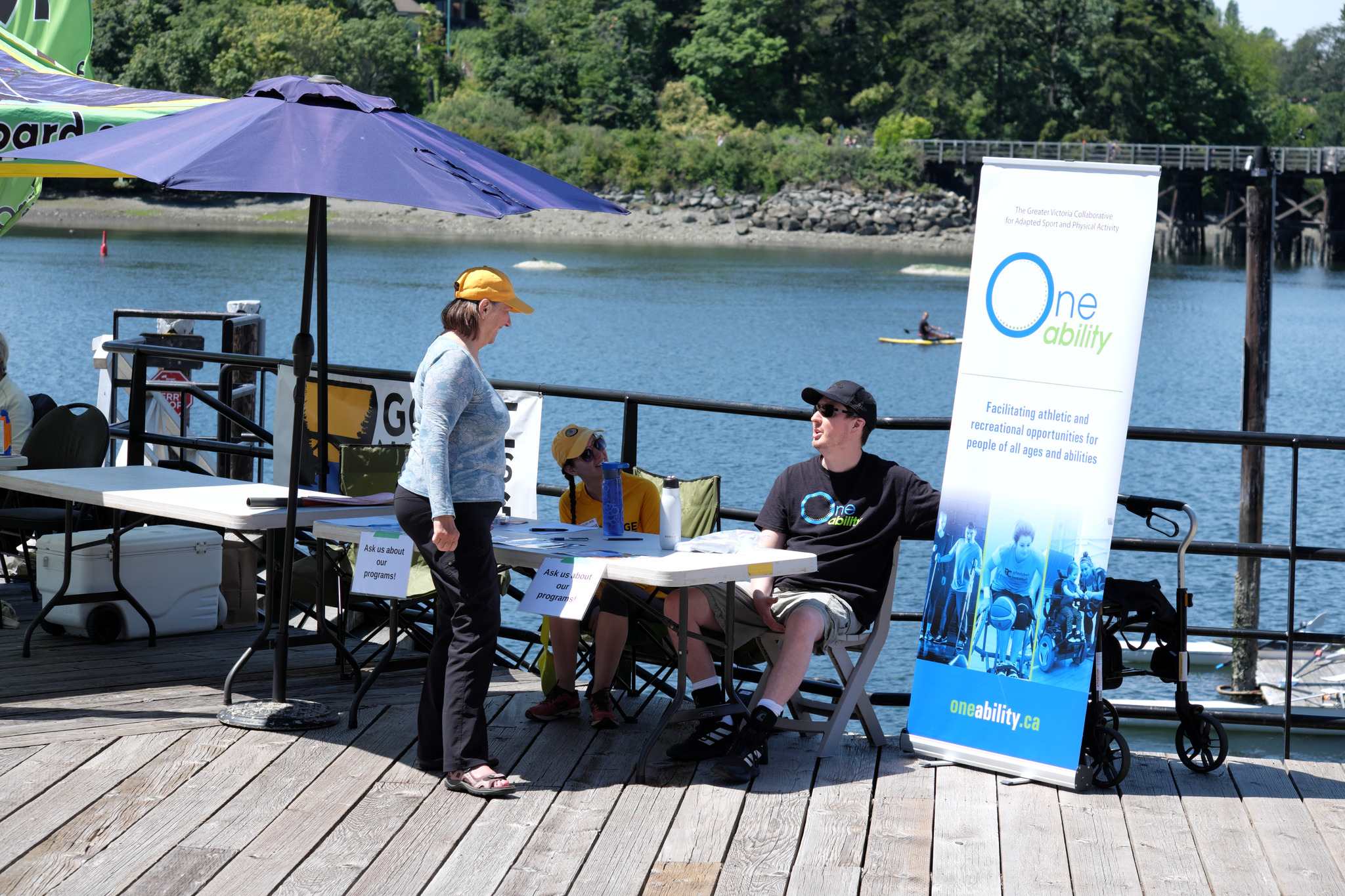 The "One Ability" desk (https://oneability.ca/). There are two people at the desk, and a large poster on the right. A woman is talking to one of the representatives. 