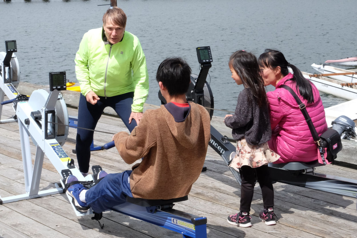 Two children get instruction from a volunteer along with their mom. One child and the mother are on rowing machines.