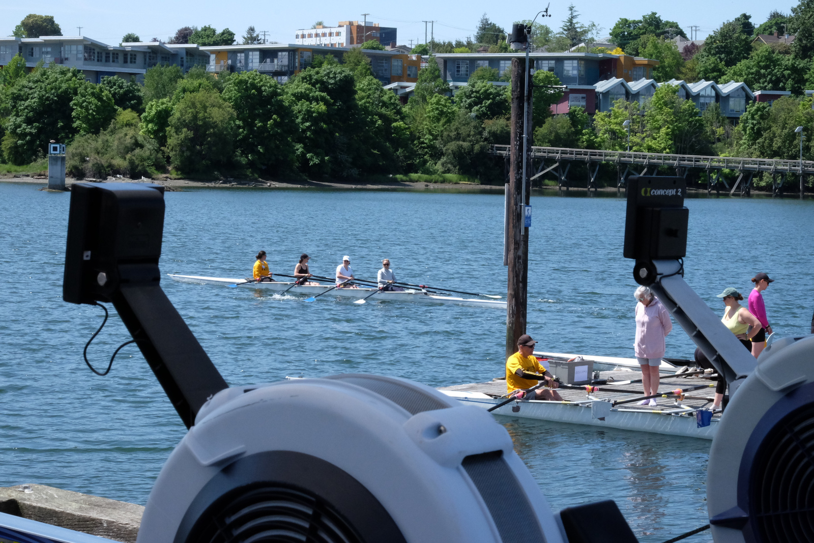 A quad on the water. In the foreground are rowing machines, another quad at the dock, and people standing.