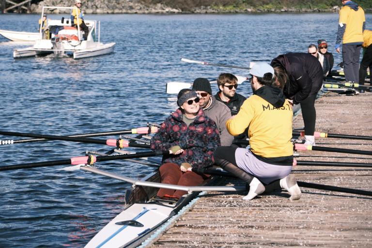 Two quads on the dock, with coach boats in the background. Coaches and volunteers are helping the participants adjust their seat and oars.