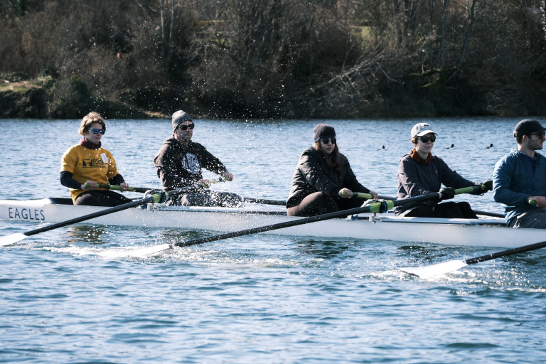 The stern five rowers of the eight on the water. Their oars are feathered (flat), and there's a spray of water visible. In the background are three evenly spaced ducks and the shore.