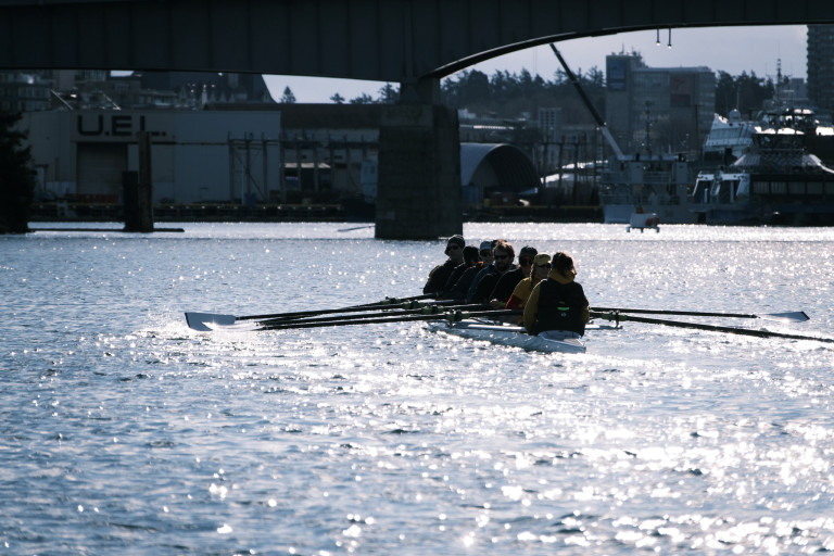 The eight heading to the Point Ellice (Bay Street) Bridge. The boat is headed into the sun, and the rowers are dark and silhouetted against the bright water.
