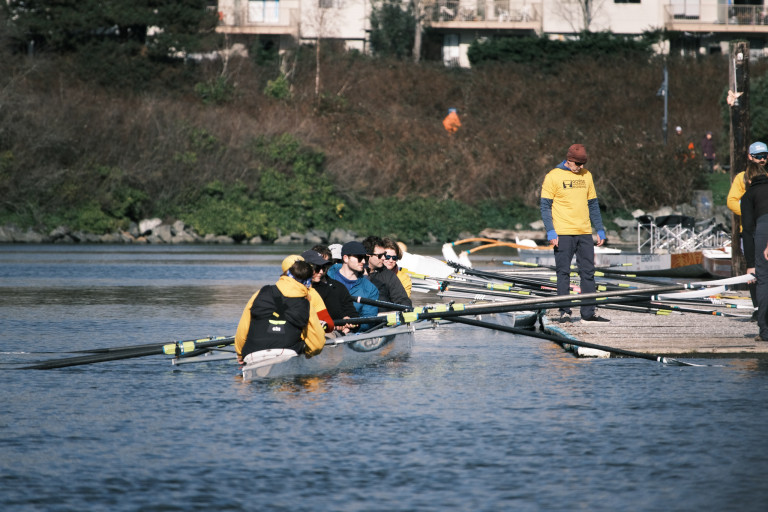 A quad coming into the dock. They are 'leaning away' from the dock so their oars will  clear. A volunteer is holding one of the oars to pull the boat in.