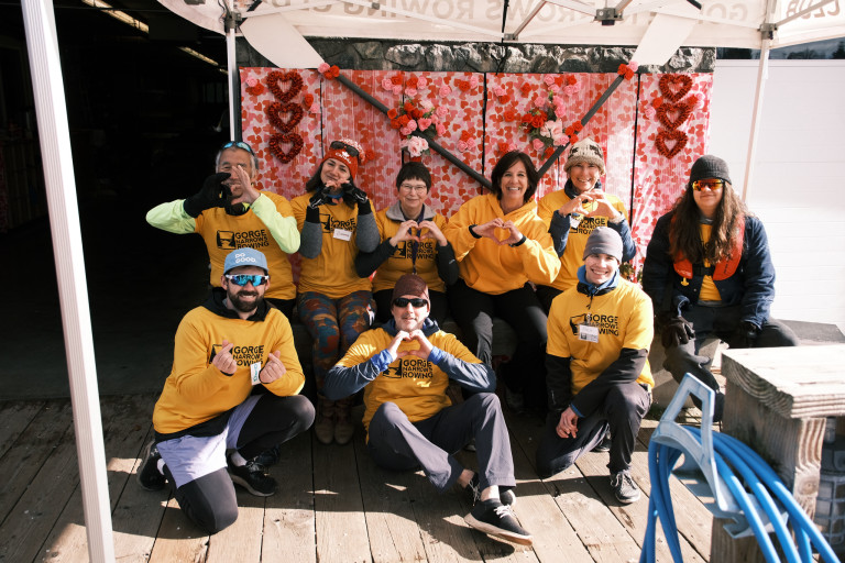 Our coaches and volunteers against a backdrop of hearts and crossed oars.