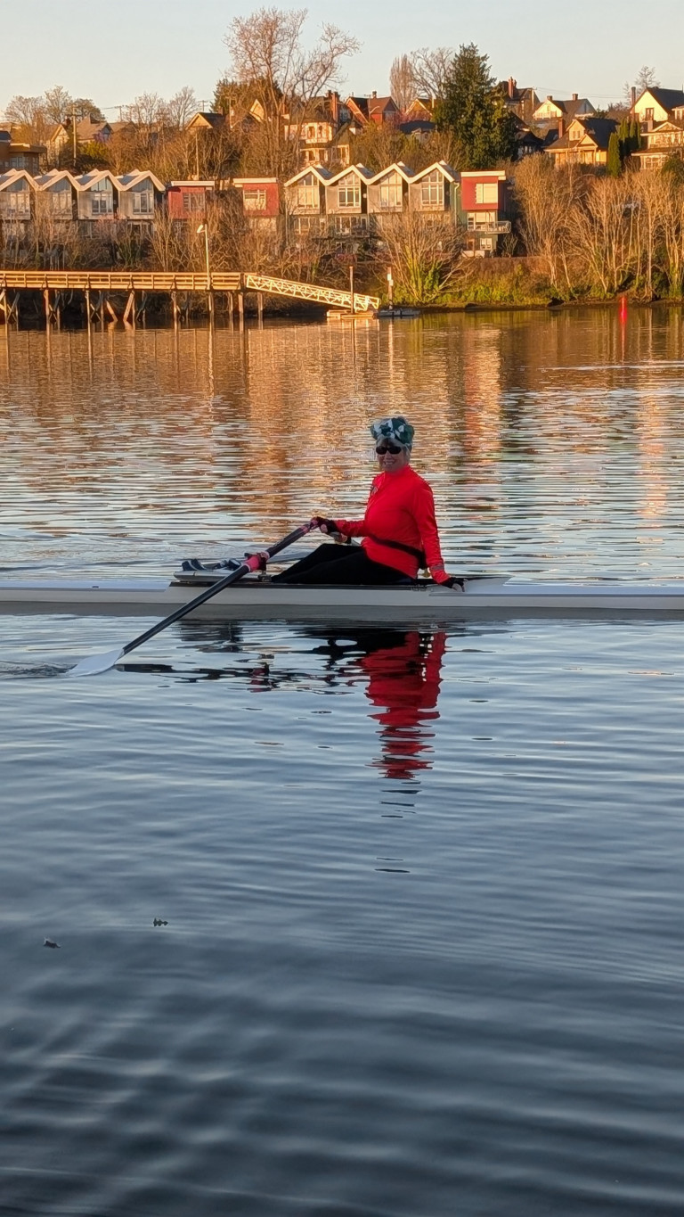 A rower in a single. In the background is the west shore of the Gorge, glowing in the early morning sunshine.