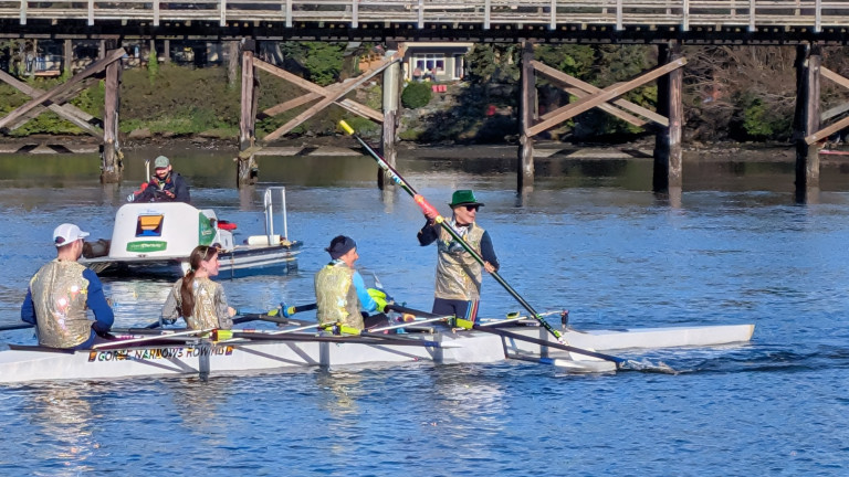 A festively dressed quad sits still as the stroke (the person in the rear-most seat) kneels facing backwards and performs  a stunt. A coach boat and the Selkirk Trestle are in the background.